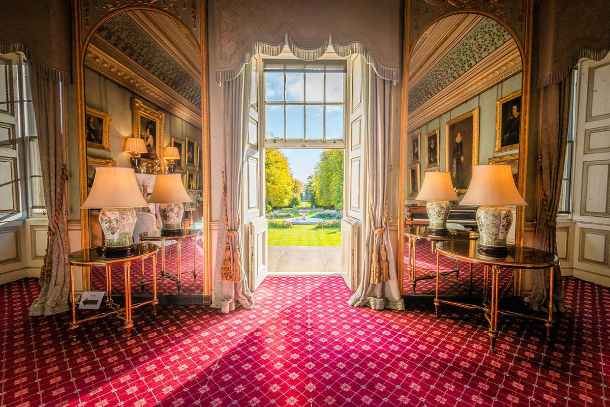 Elegant drawing room with gold-framed mirrors, decorative lamps, and large open doors leading to landscaped gardens