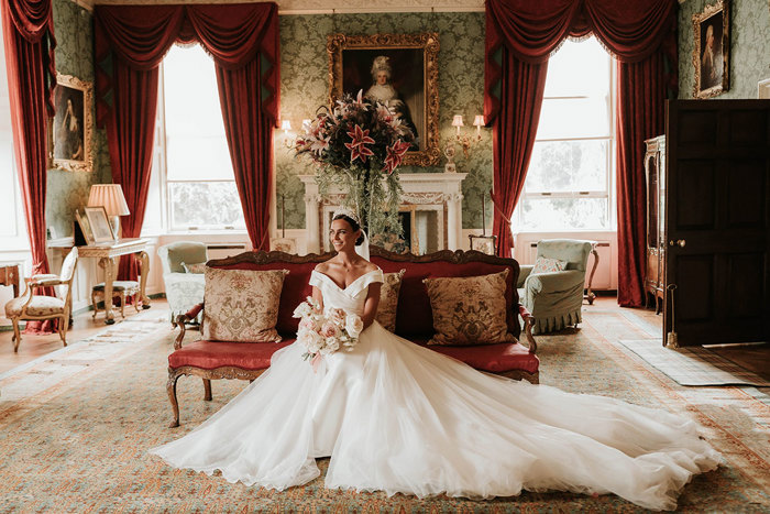 A bride sits on an old fashioned couch in a grand room with green walls and red curtains. She is holding a bouquet of pale flowers and wearing a white crown