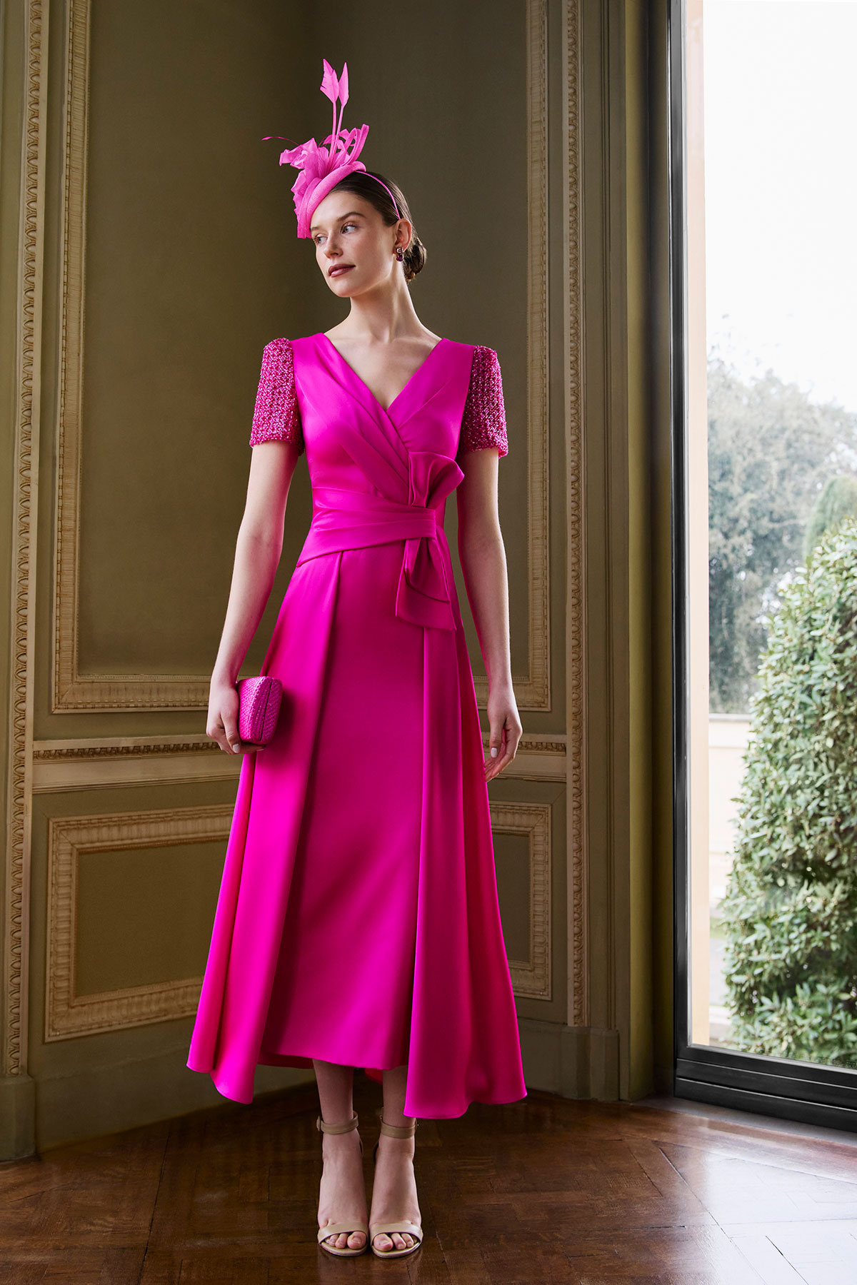 Mother of the bride outfit in a bright fuchsia midi dress with embellished sleeves, matching headpiece and clutch bag.