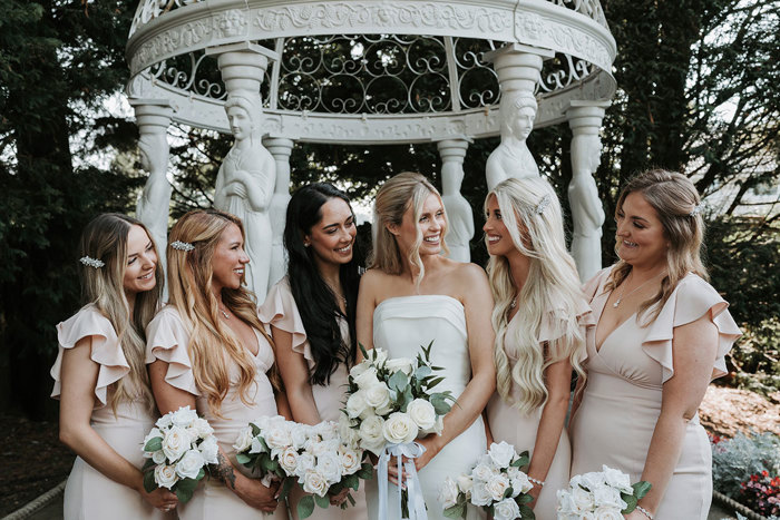 a line of bridesmaids and a bride posing for a photo in garden of Busby Hotel in front of white ornamental pergola