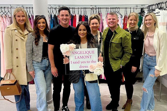 A person holding a sign that reads 'Angelique Lamont I said yes to the dress' surrounded by seven other smiling people standing in a row against a rail of colourful dresses