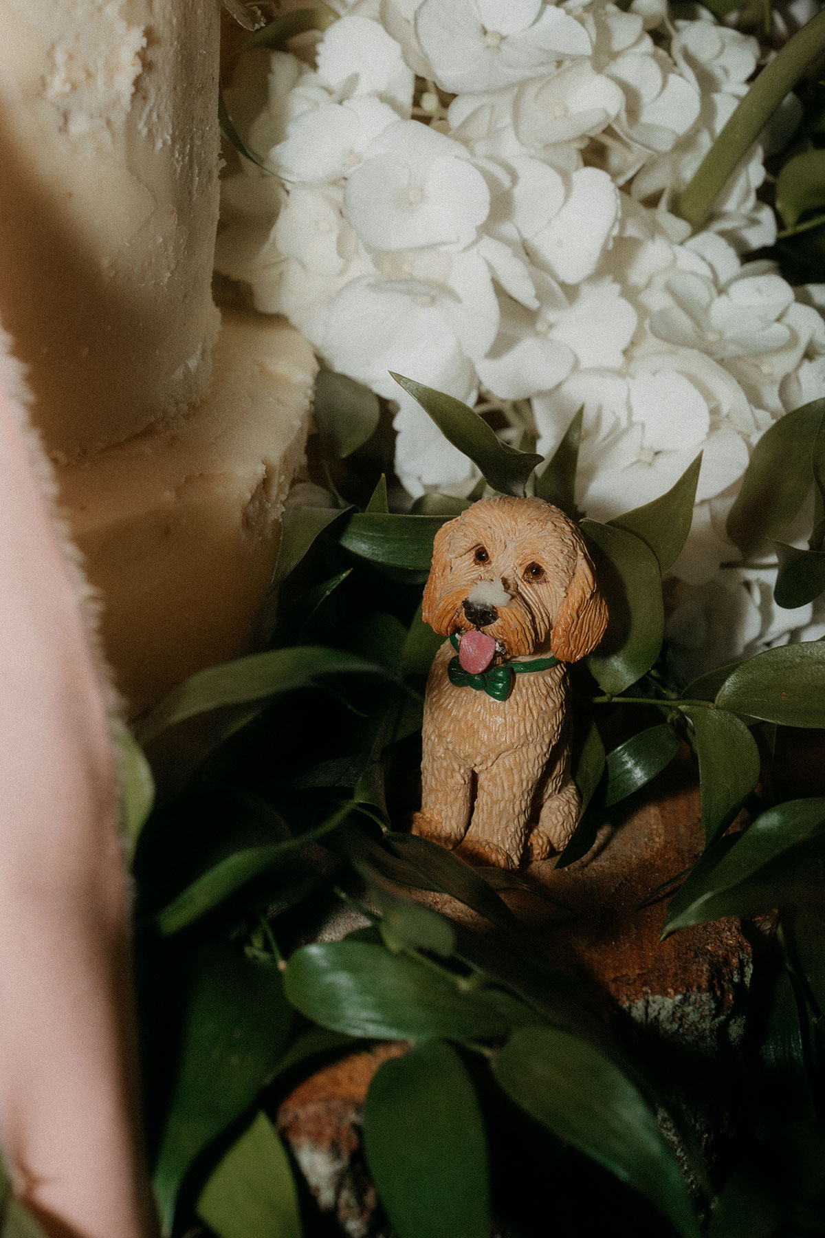 Close-up of dog figurine wedding cake topper nestled among greenery and white flowers at rustic barn wedding reception