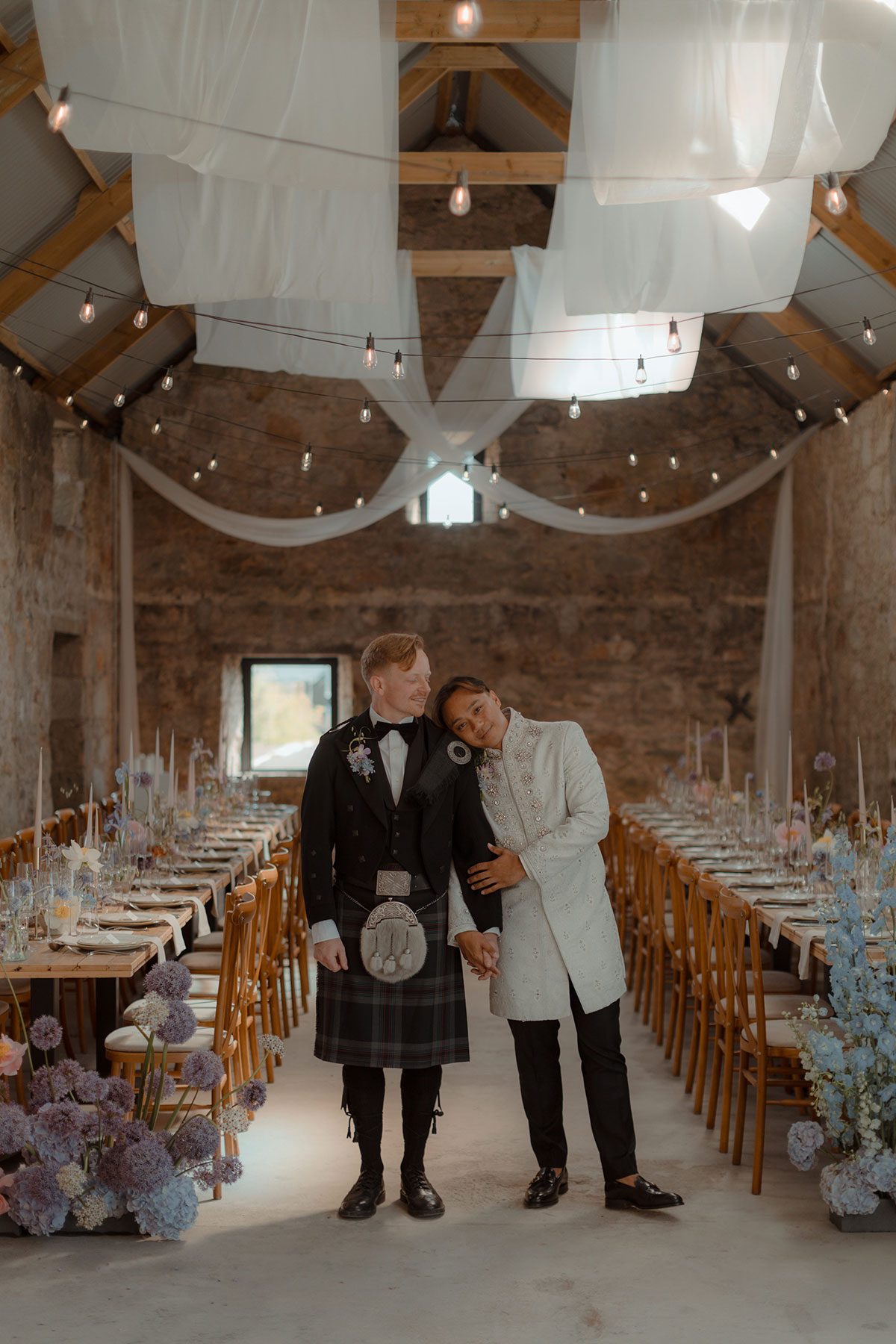 Same-sex couple standing in decorated barn with long banquet tables and soft ceiling draping at Falkirk farm wedding