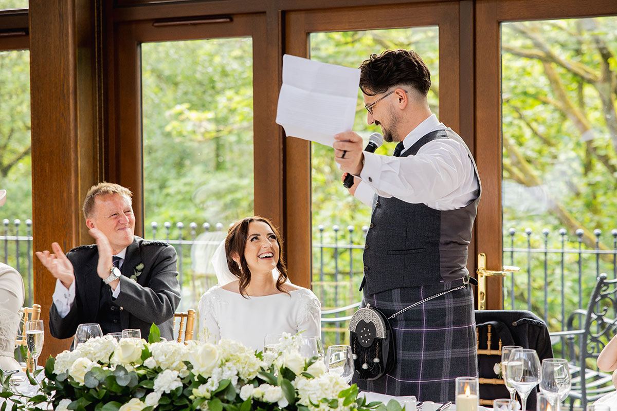 Groom gives a speech in front of large windows while the bride smiles up at him and guests applaud at the top table decorated with white flowers