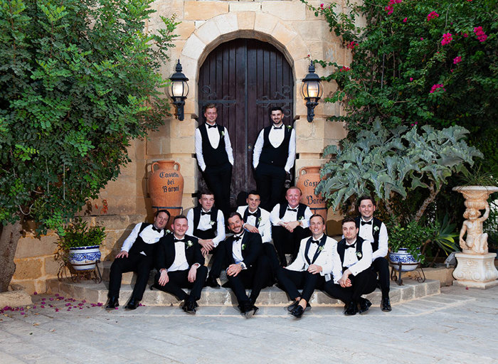 a group of men wearing smart suits, waistcoats and bow ties posing outside a stone doorway. Some are sitting on stone steps; some are standing.