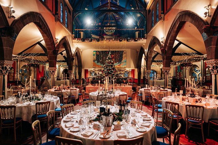 dinner celebration is set up in a large space with at least six round fully set dining tables, complete with candelabras and a tall fully decorated Christmas tree at the head of the room