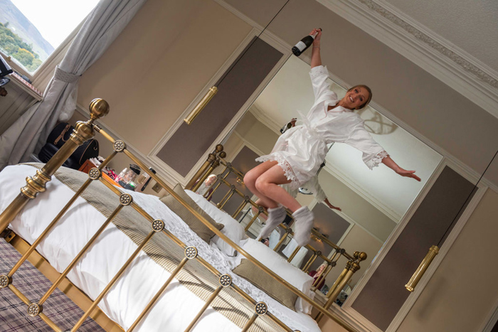A Person Jumping On A Bed While Holding A Bottle Of Asti