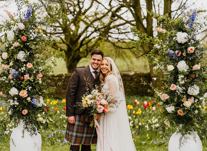 a bride and groom in a garden setting that is carpeted with colourful tulips. They are flanked by two large white urns that are filled with a tall column arrangement of pastel coloured flowers