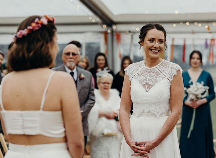 a smiling bride wearing a fine lace top and flowing skirt clasps her hands as a second bride wearing a flower crown and white crop top faces her. There are other wedding guests in the background looking on in a marquee setting with colourful paper streamers hanging from strings that run across the marquee