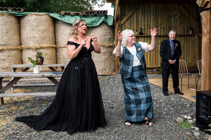 A Bride Wearing A Black Wedding Dress And Person Wearing Tartan Waistcoat And Skirt Clap Outside A Wooden Building With Bales Of Hay In Background