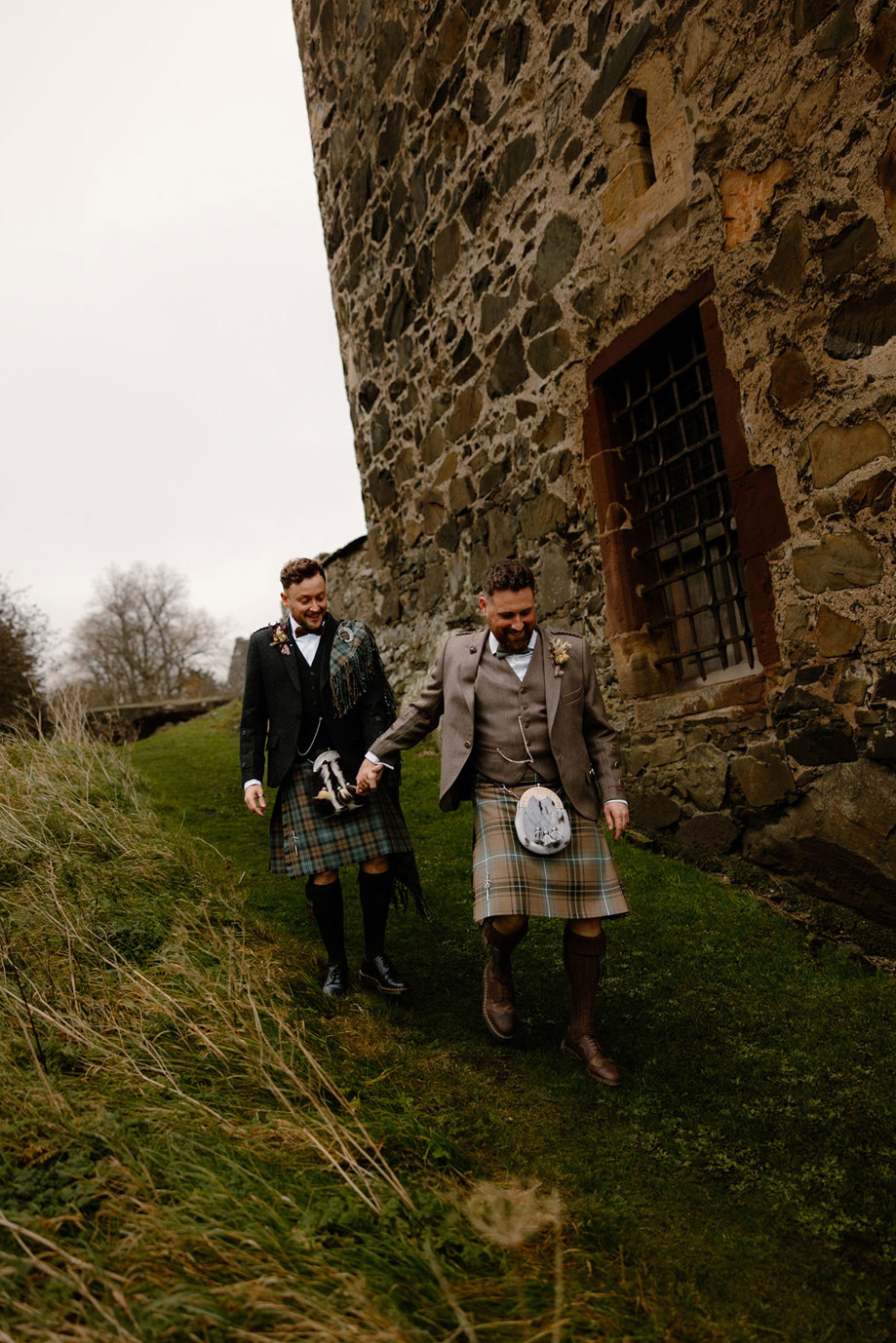 two grooms holding hands walking down a small grassy slope outside the old stone walls of Neidpath Castle