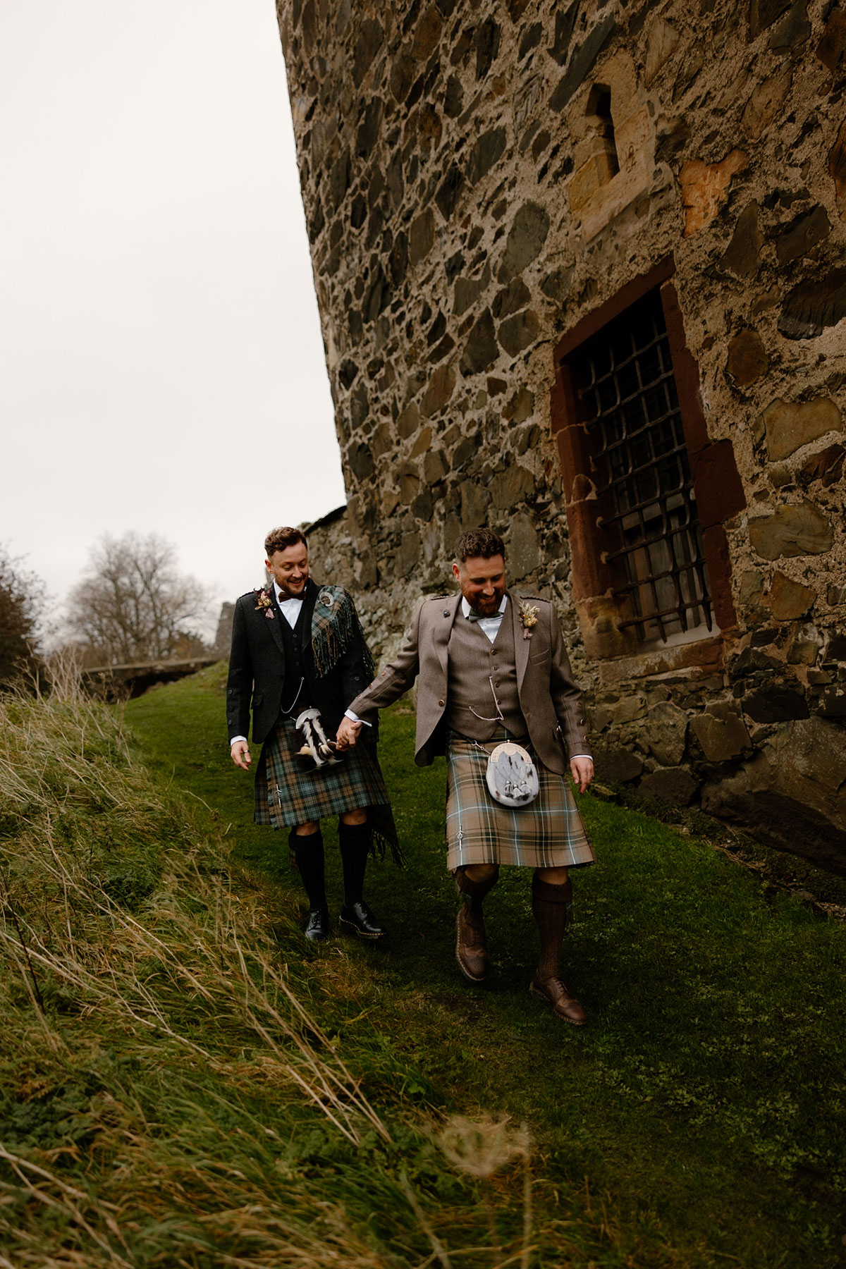 two grooms holding hands walking down a small grassy slope outside the old stone walls of Neidpath Castle