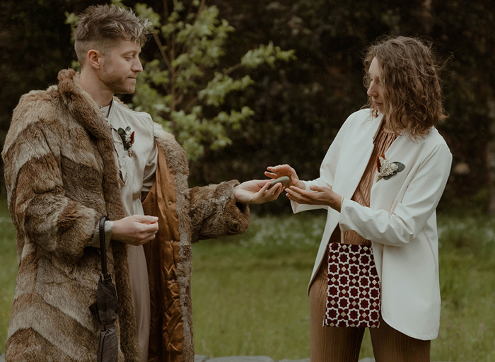 a person wearing a brown fur style jacket passing an oathing stone to a person wearing a white jacket. They are outdoors in a garden setting