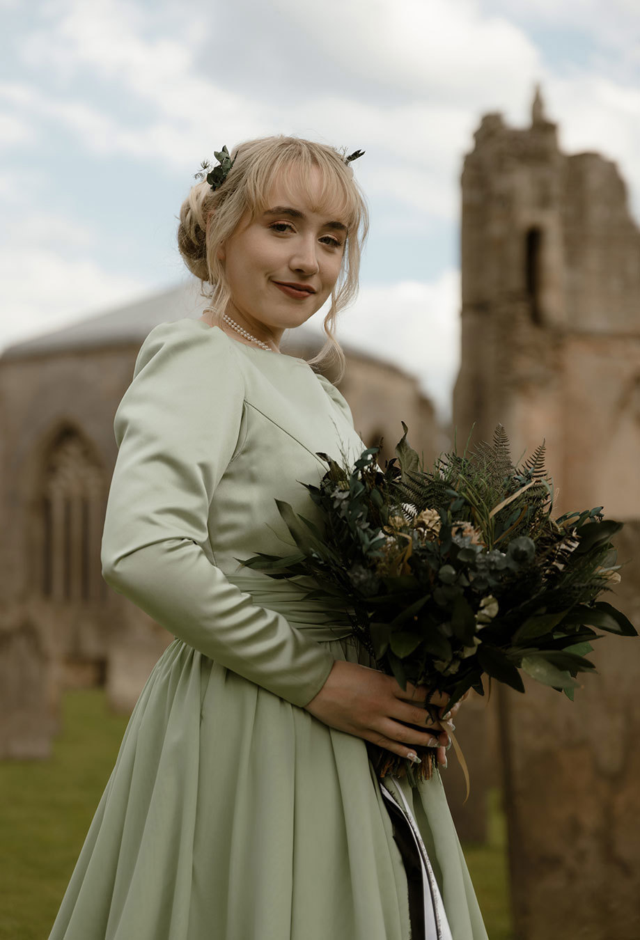 a softly smiling bride wearing a light green dress and holding a green foliage bouquet. There is a church in the background