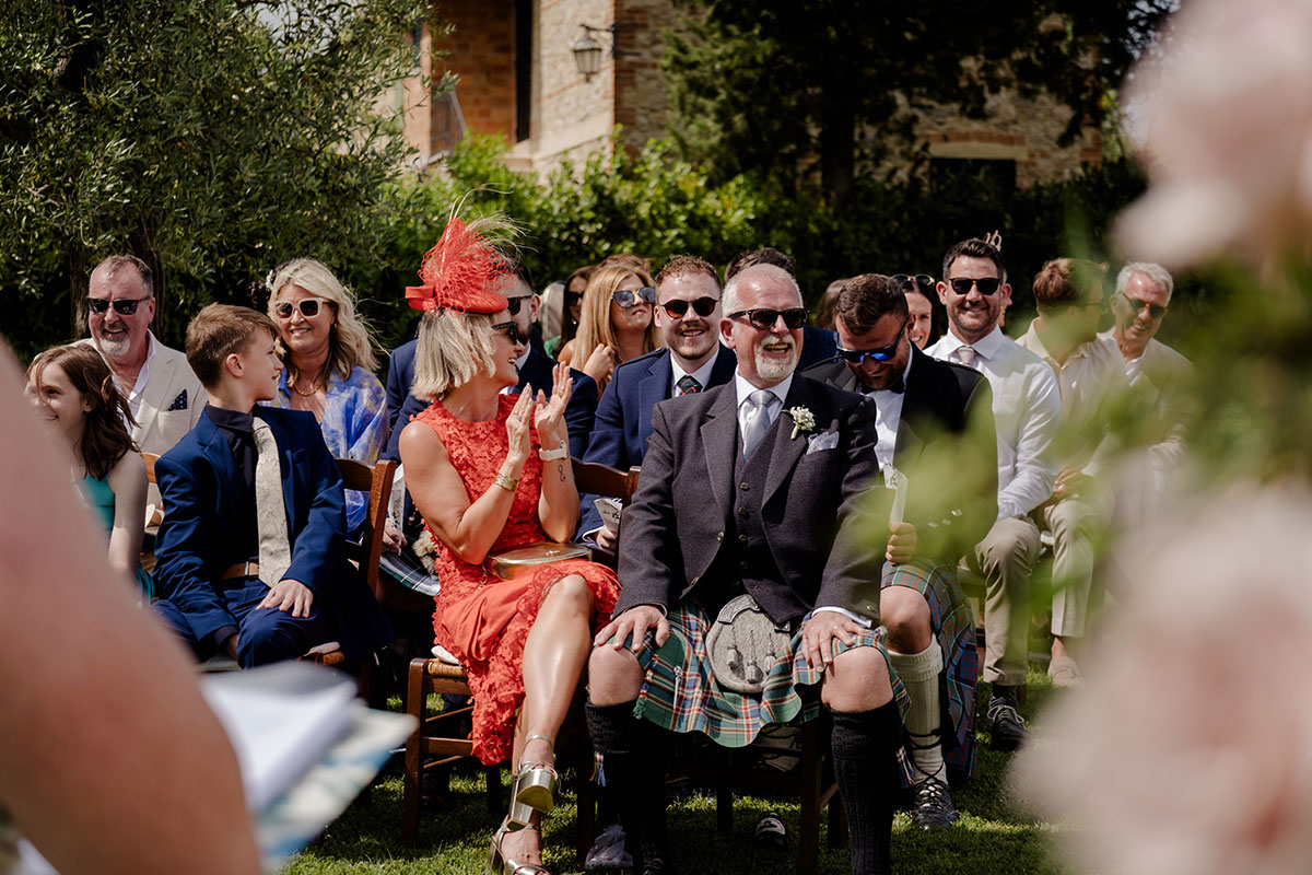 rows of guests sit on wooden chairs outside, wearing sunglasses and a range of formal outfits from suits and ties, kilt outfits, and dresses 
