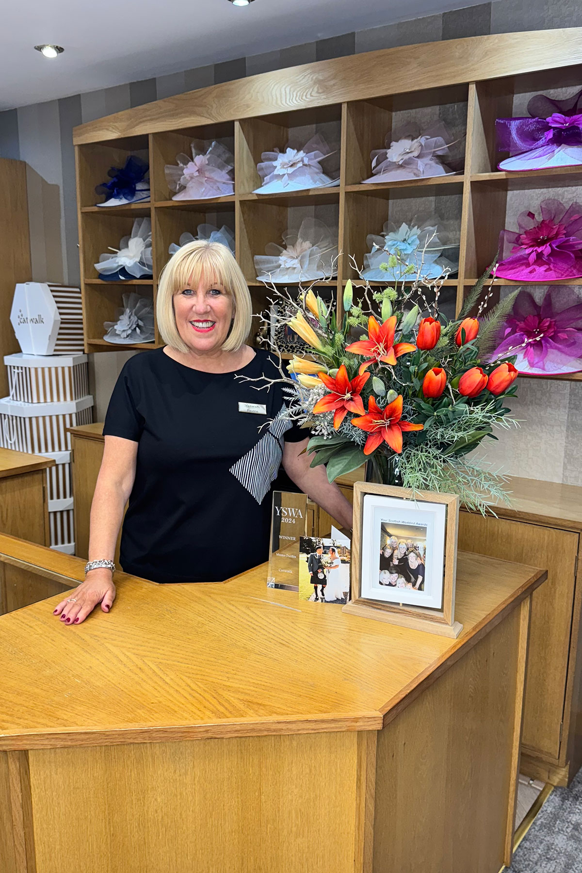 Bridal boutique owner standing behind counter with bright floral arrangement and award display, inside Scottish mother-of-the-bride shop
