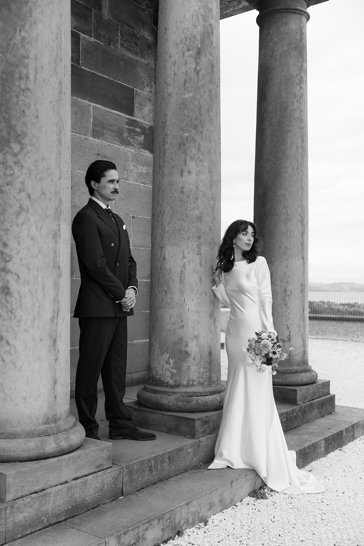 Bride and groom posing between stone pillars at the City Observatory on Calton Hill during their Edinburgh wedding.