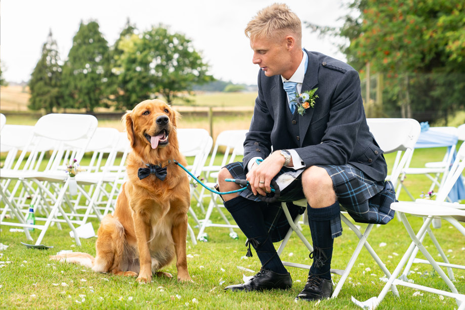 A man in a kilt sits on a chair next to a golden retriever wearing a bowtie