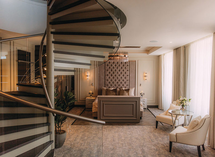 A luxurious bedroom in a hotel with a spiral staircase leading upstairs on the left, a kingsized bed with beige and brown furnishings and two white armchairs