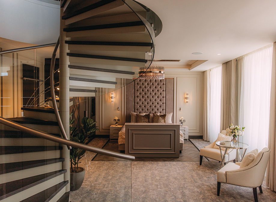 A luxurious bedroom in a hotel with a spiral staircase leading upstairs on the left, a kingsized bed with beige and brown furnishings and two white armchairs