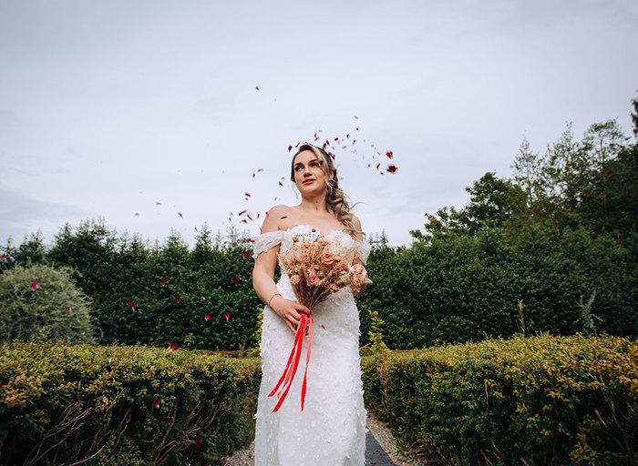 bride in an off the shoulder wedding dress holds a dried bouquet of flowers and has petals falling around her face