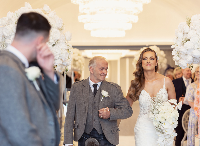 a groom wipes away a tear as a bride walks towards him on the arm of a man wearing a kilt