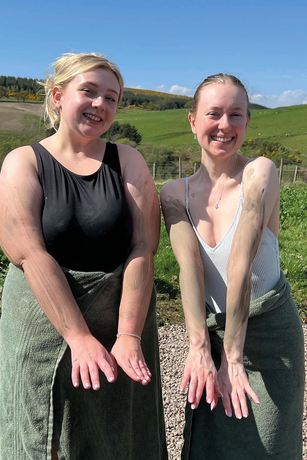 two blonde women in swimsuits with green towels around their waists smile as they hold out their arms, all of which have been lathered in a grey clay mud