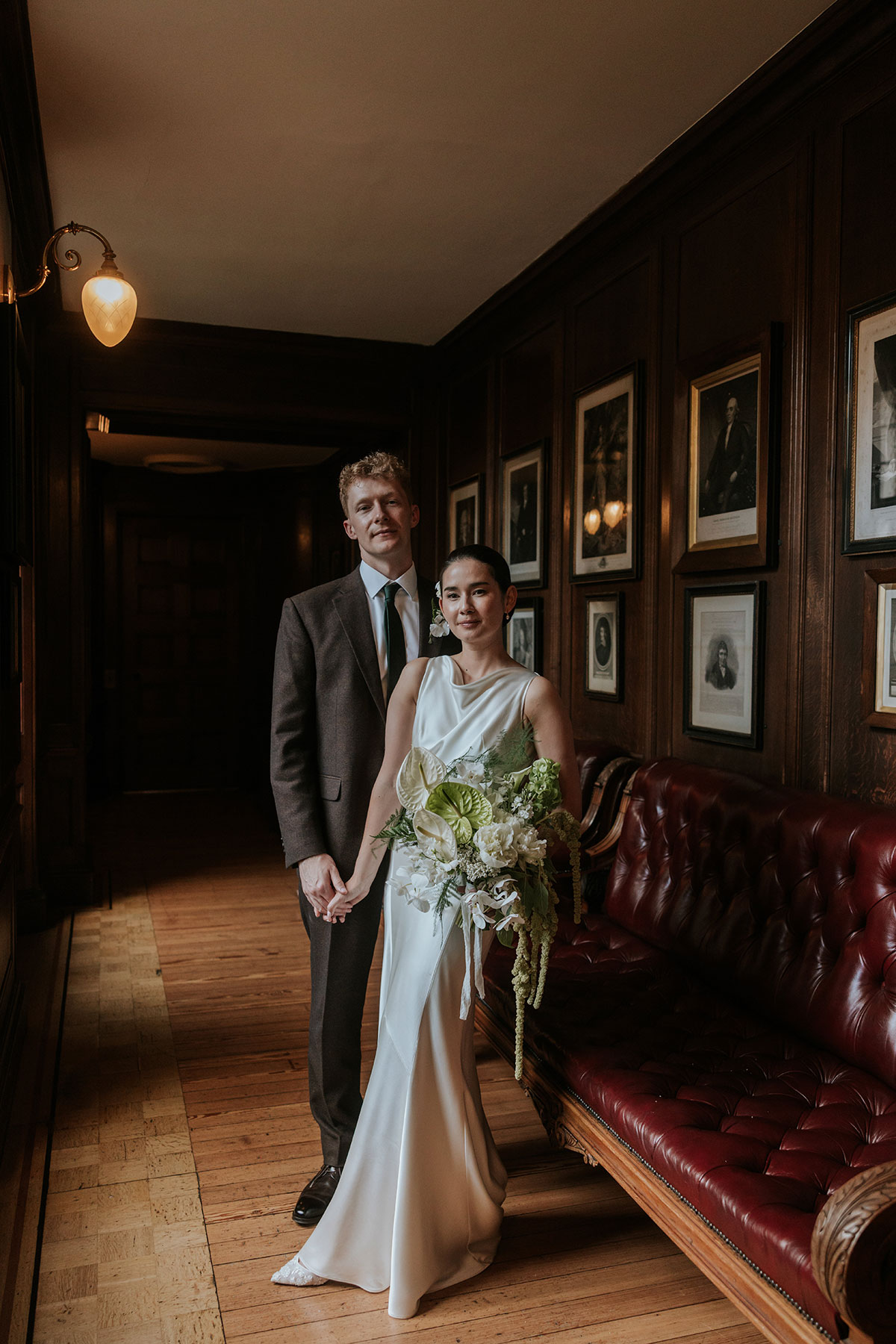 Bride and groom standing hand in hand beside a red leather couch, bride holding bouquet.