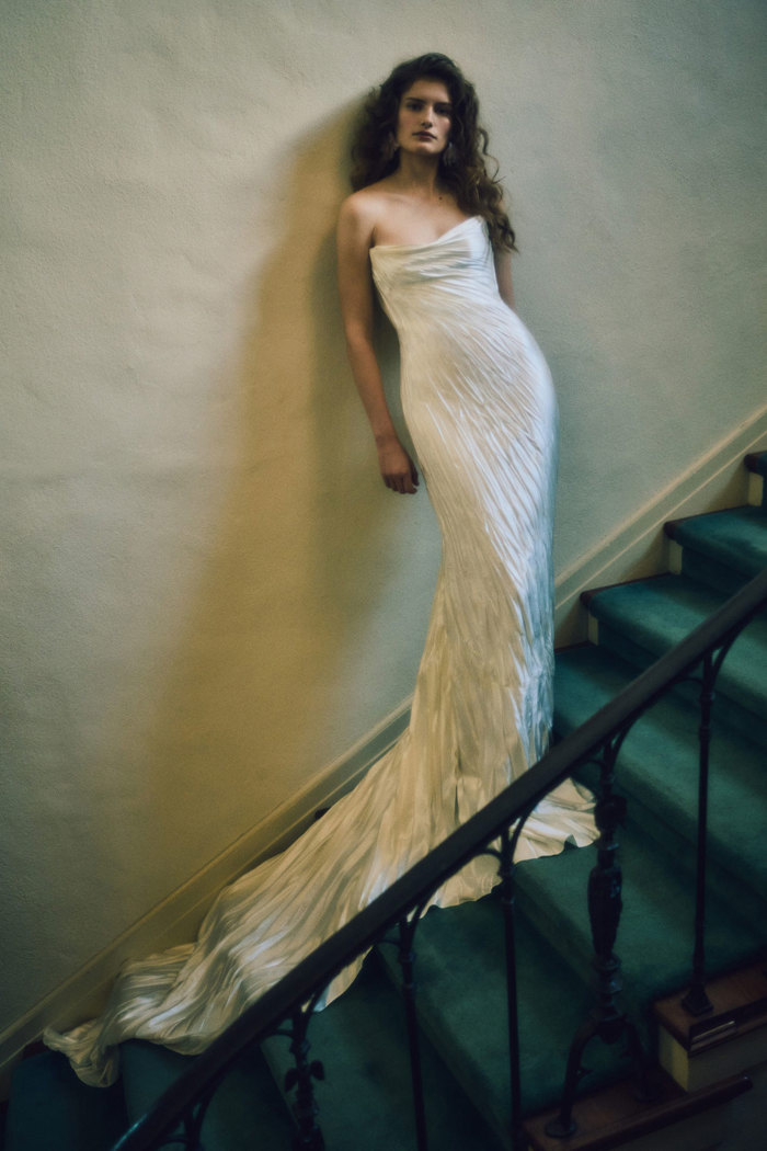 a darkened image of a brunette model stood on a green carpeted staircase wearing a strapless wedding dress with crinkled pleated and a boned bodice