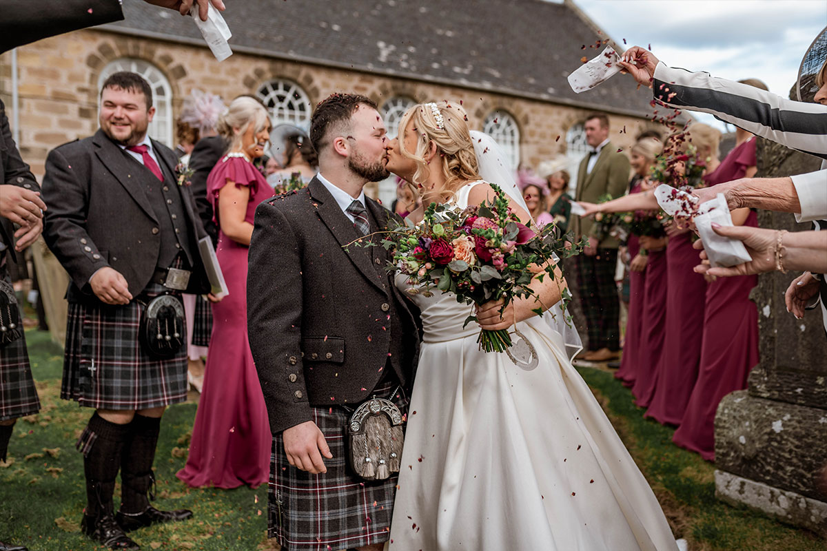 A bride and groom kiss while their guests throw confetti over them