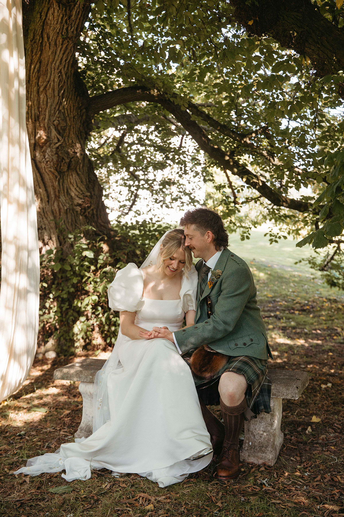 Bride and groom seated beneath large tree for relaxed portraits at Highland farmhouse wedding in Nethy Bridge