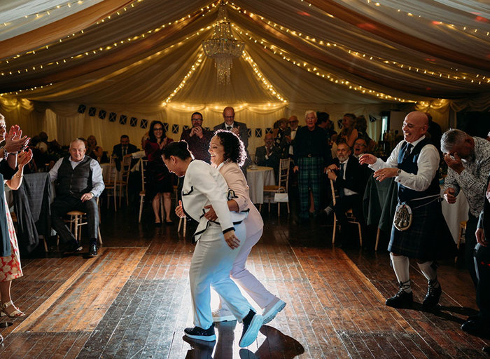 two people wearing suits dancing on a wooden floor in a marquee as guests look on