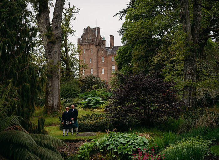 two grooms in lush green gardens with Brodick Castle in the background