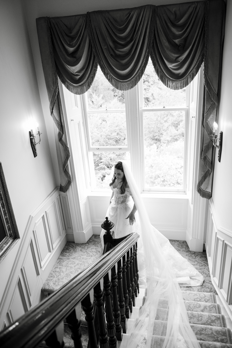 Black-and-white portrait of bride on the staircase at Crossbasket Castle with her cathedral-length veil trailing behind her.