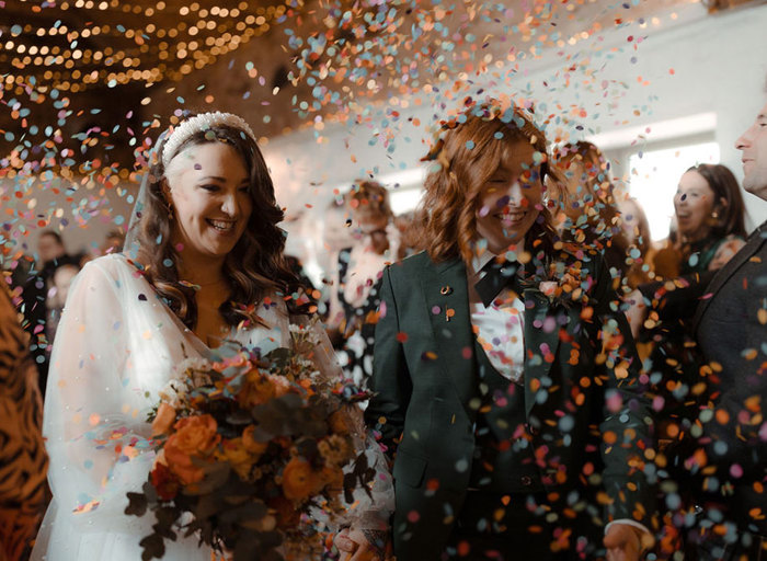 two brides being showered in colourful confetti with blurred out guests in the background