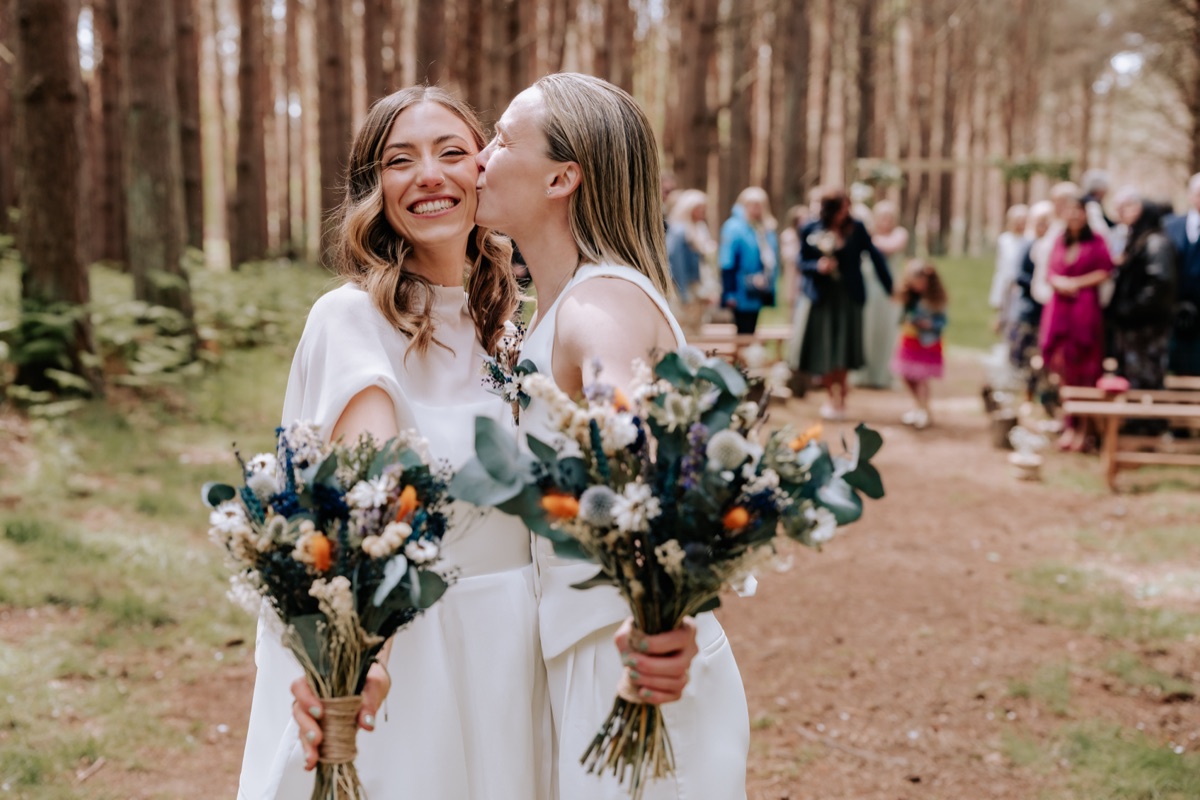 Hannah and Donna holding rustic wedding bouquets as one bride kisses the other on the cheek at Harvest Moon.