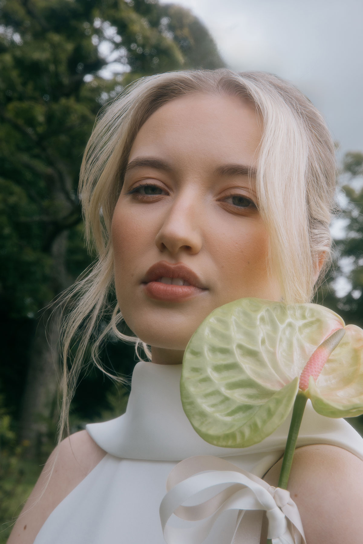 A close-up portrait of a bride holding a green anthurium flower near her face, with soft makeup and blurred outdoor greenery behind her.