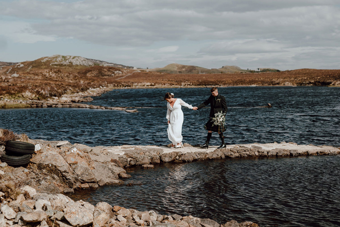 Newlyweds hold hands as they cross strip of land near water