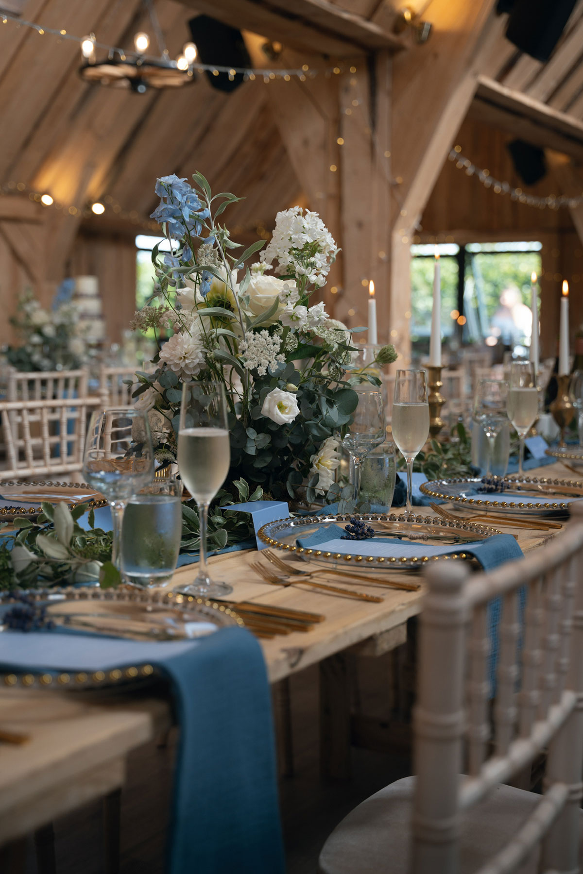 Wedding reception table styled with blue runners, gold-rimmed charger plates, candles and tall floral centrepieces inside a wooden barn venue