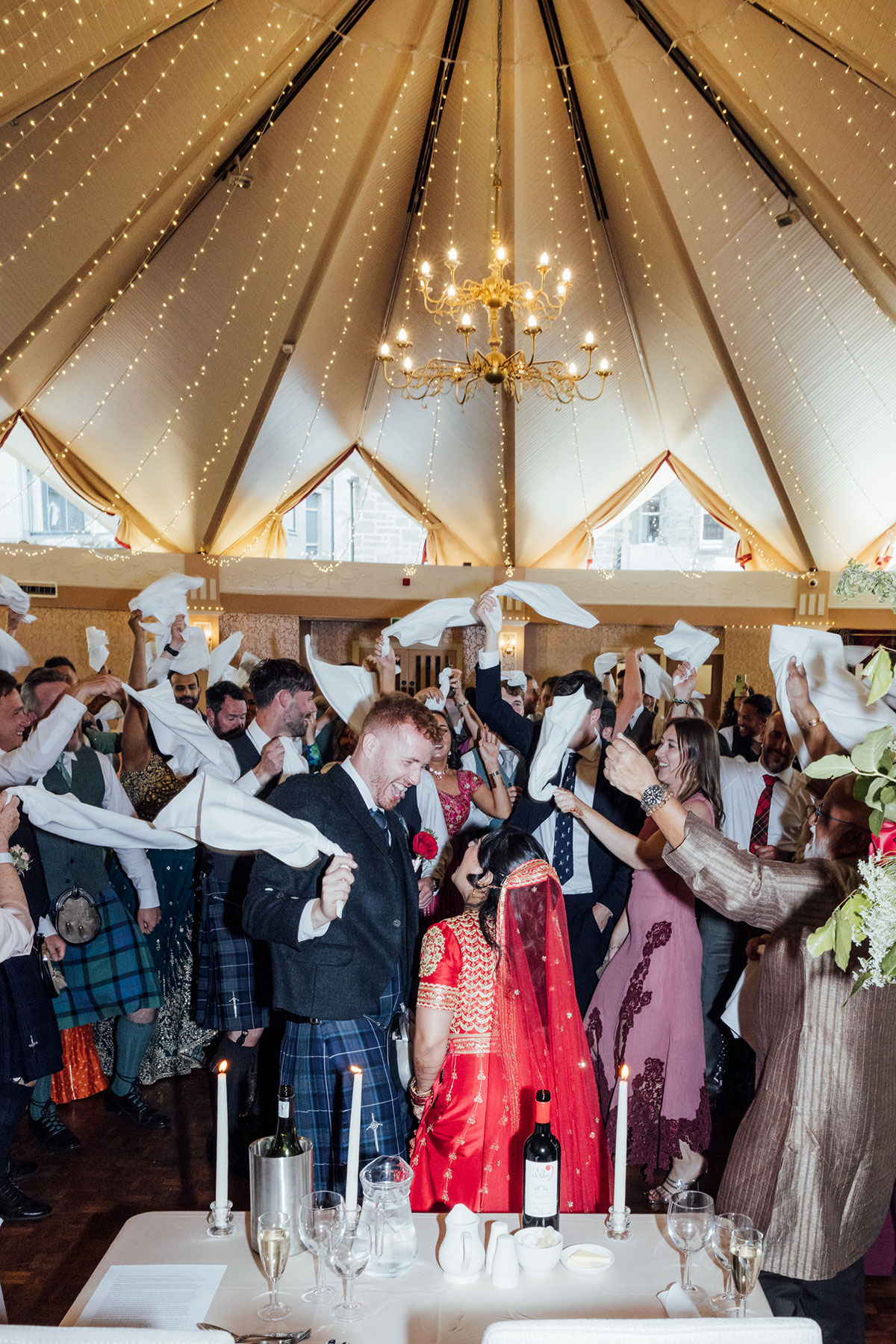 Bride and groom dancing together as guests wave napkins during their lively Scottish and Indian wedding reception