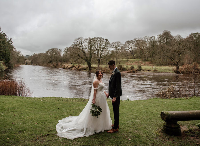 A bride and groom stand on grass in front of a river 