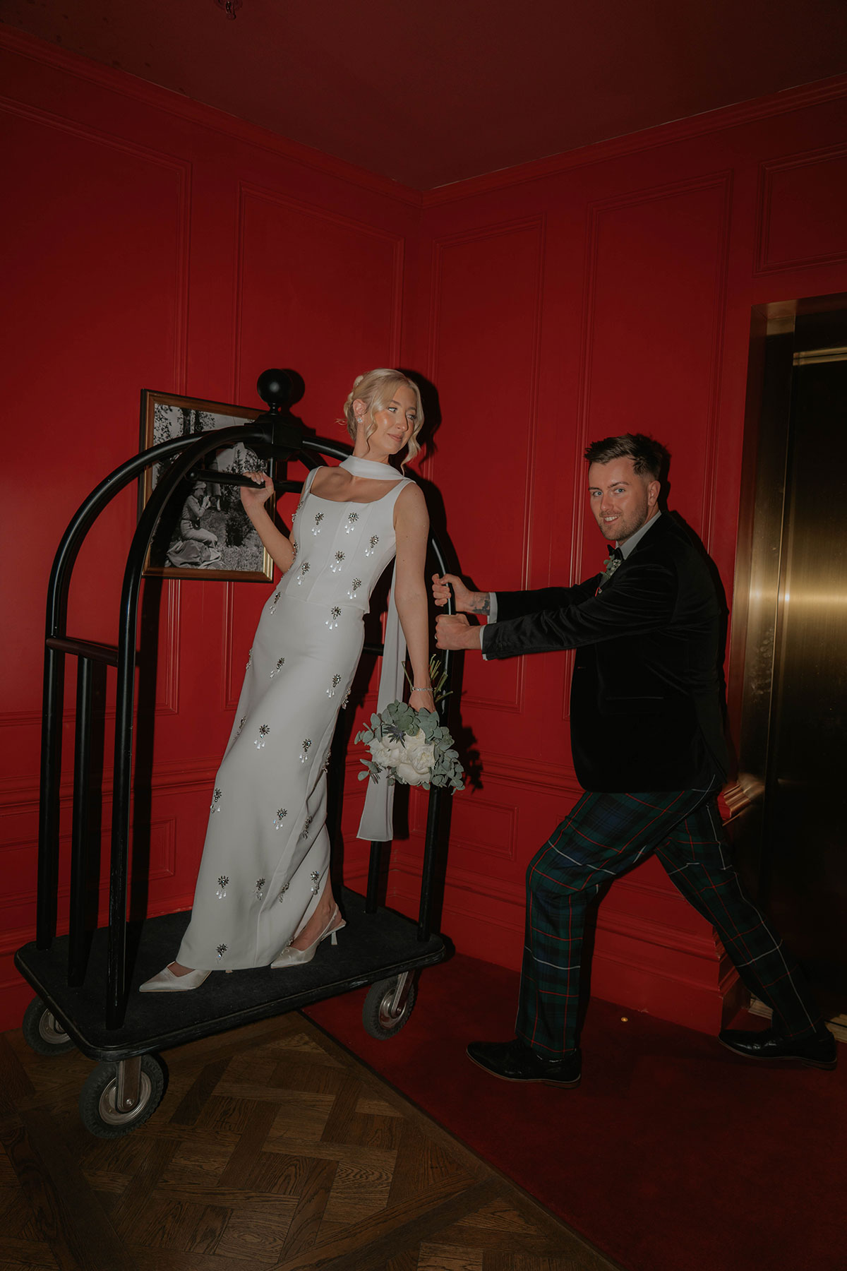 Bride and groom having fun with a hotel luggage trolley against a bold red backdrop