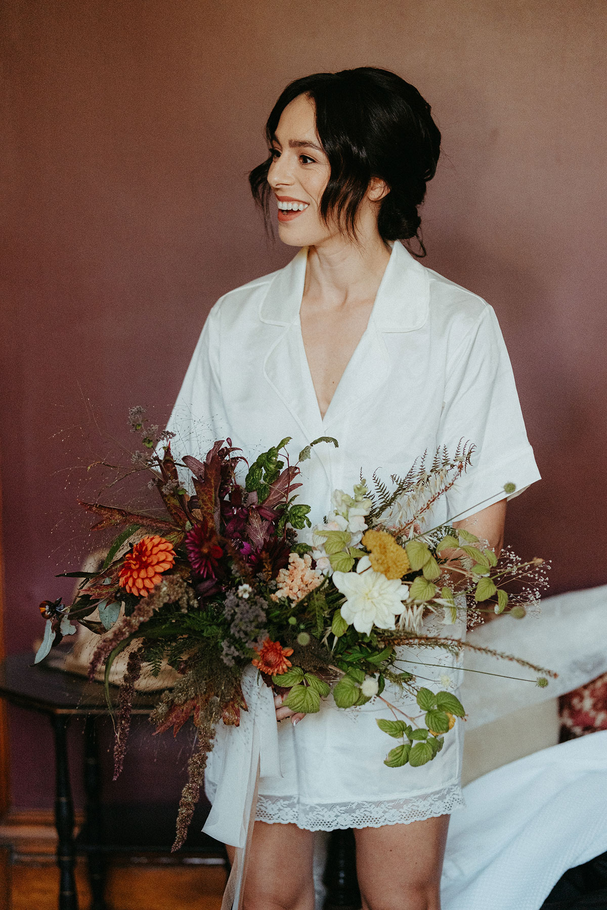 Bride in white bridal pyjamas holds her colourful autumn bouquet while getting ready ahead of her Cambo Country House wedding in Fife.