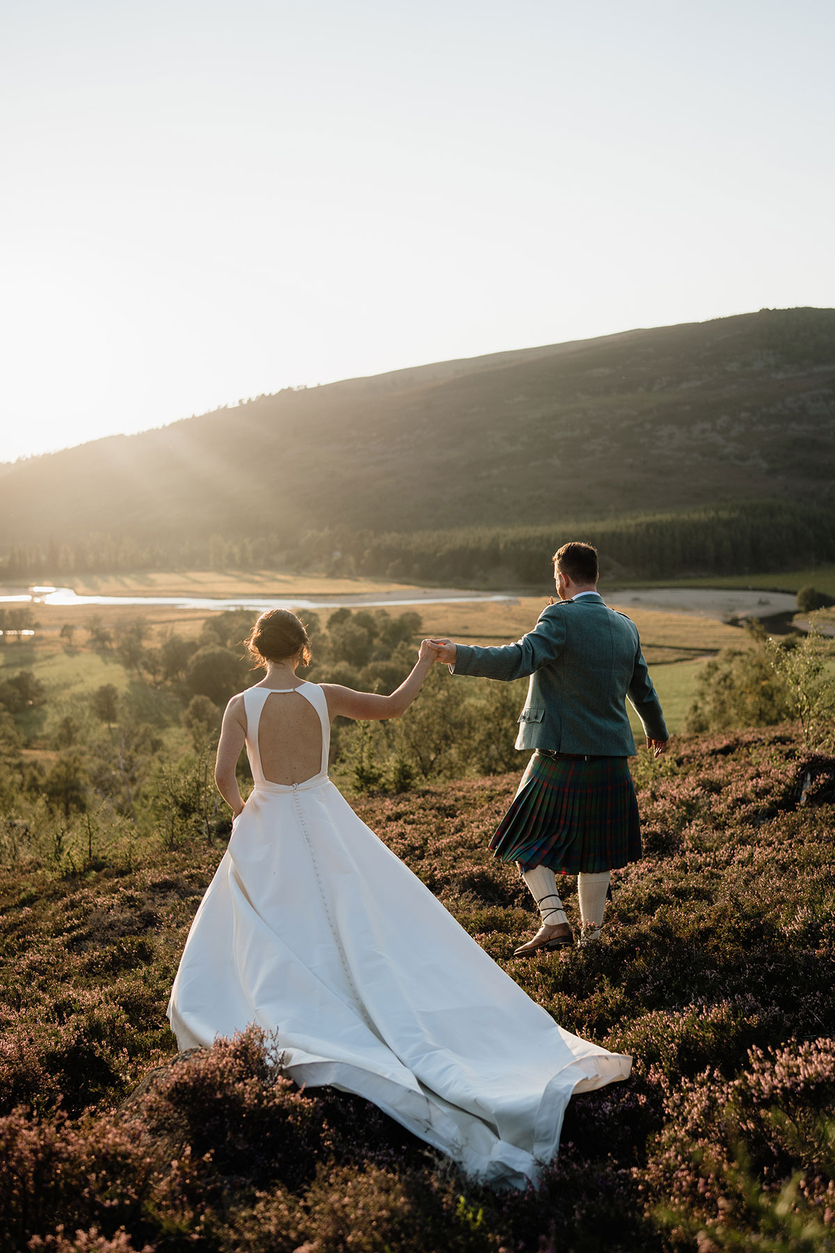 Bride and groom holding hands on a heather-covered hillside overlooking the Cairngorms during golden hour wedding portraits