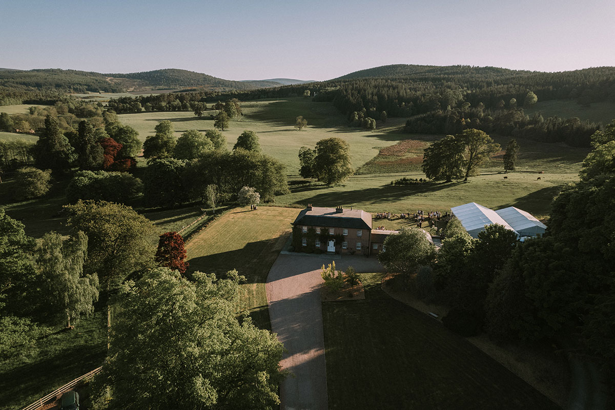 An aerial shot of a country house and marquee surrounded by rolling countryside and mature trees.