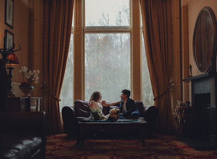 A couple sit facing each other on a couch in a warm toned room with a large widow and dark yellow curtains