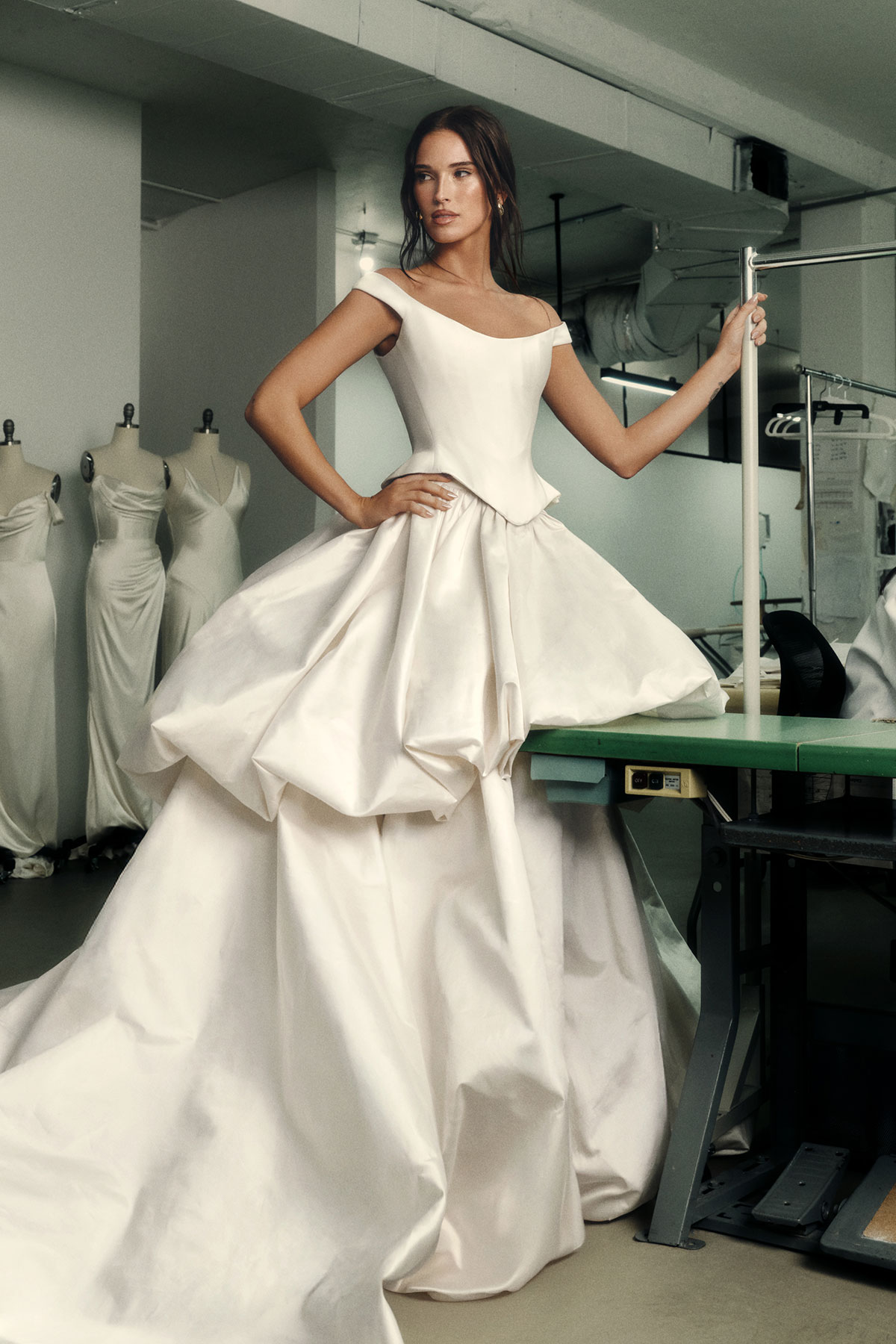 Model wearing a structured off-the-shoulder wedding dress with sculptural bodice and dramatic tiered skirt, photographed inside a bridal atelier with dress forms in the background