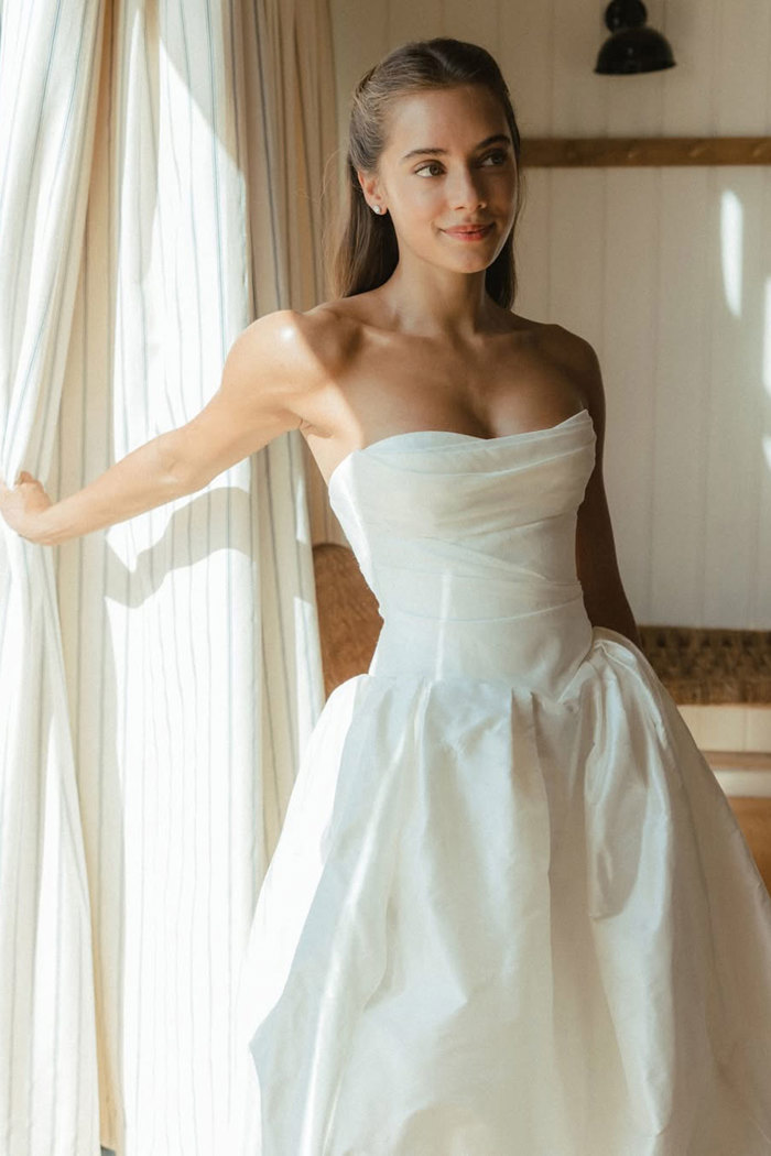 brunette woman with hair half up half down wears a floaty strapless wedding dress while reaching an arm out and holding onto the curtain behind her