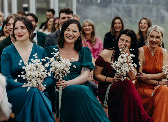 four bridesmaids wearing dresses in teal, forest green, orange and red each carrying a posy of daisies. They are laughing and there are other guests in the background