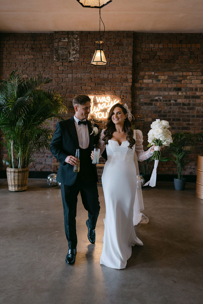 A bride in a short white dress and groom in smart suit walking in front of a brick wall with neon sign and plants behind them